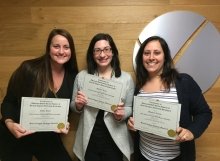 group of 3 recieve their certification in front of their company logo in the lobby